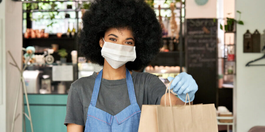 African American female cafe worker wearing face mask holding takeaway food bag. african american waitress offering takeout to customer after successfully using a takeout pos system