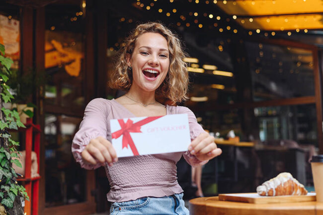 Young happy smiling woman in casual clothes sitting alone at table in coffee shop cafe eat breakfast hold gift certificate coupon voucher card for store relaxing in restaurant during free time indoors
