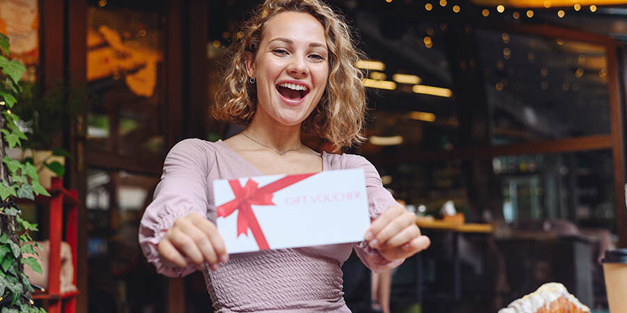 Young happy smiling woman in casual clothes sitting alone at table in coffee shop cafe eat breakfast hold gift certificate coupon voucher card for store relaxing in restaurant during free time indoor Young happy smiling woman in casual clothes sitting alone at table in coffee shop cafe eat breakfast hold gift certificate coupon voucher card for store relaxing in restaurant during free time indoors
