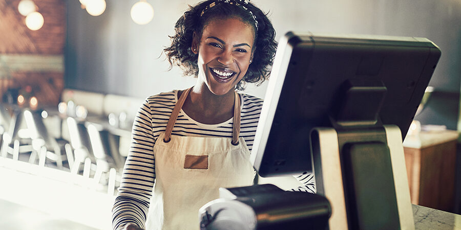 Young waitress wearing an apron standing by a point of sale terminal and laughing while working in a trendy restaurant