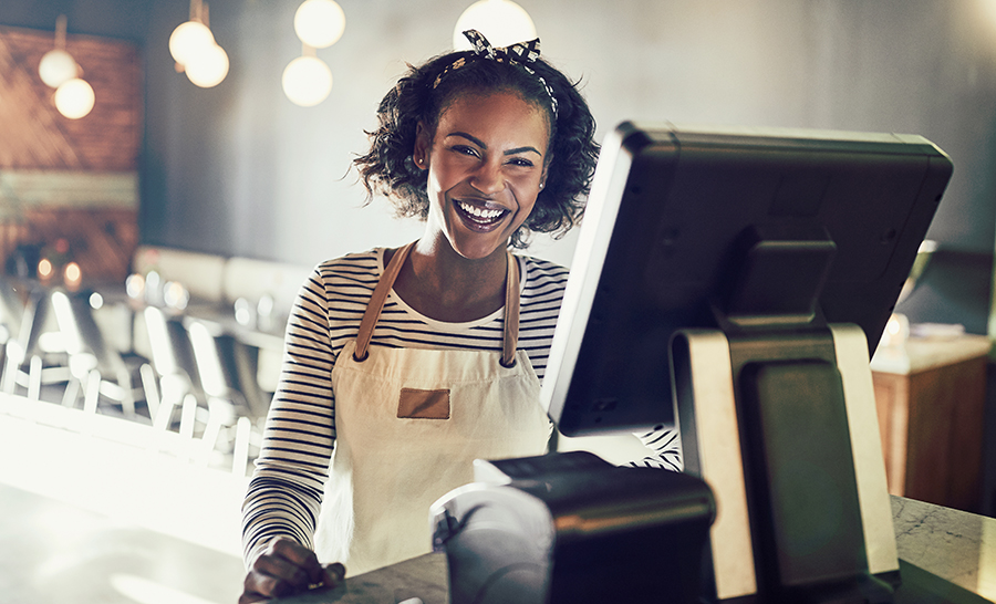 Young waitress wearing an apron standing by a point of sale terminal and laughing while working in a trendy restaurant