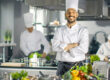 Famous Chef of a Big Restaurant Crosses Arms and Smiles in a Modern Kitchen. His Staff in Working in the Background.