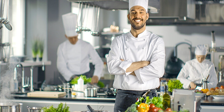 Famous Chef of a Big Restaurant Crosses Arms and Smiles in a Mod Famous Chef of a Big Restaurant Crosses Arms and Smiles in a Modern Kitchen. His Staff in Working in the Background.