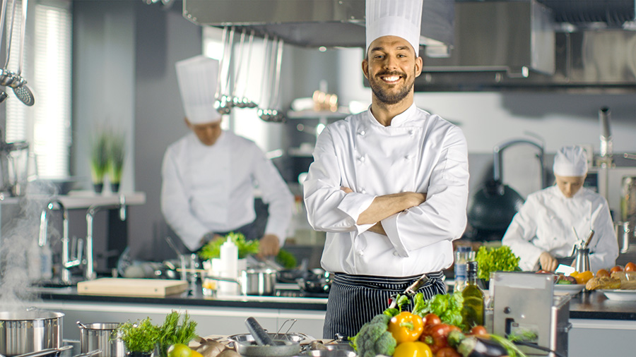 Famous Chef of a Big Restaurant Crosses Arms and Smiles in a Modern Kitchen. His Staff in Working in the Background.