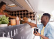 street sale, payment and people concept - happy african american young man buying wok and paying with dollar money at food truck